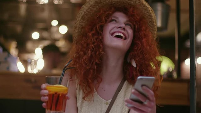 Smiling young attractive pretty woman with red hair in hat sitting in cafe use phone drinks a cocktail happy fashion girl connection summer portrait caucasian smartphone lady close up slow motion