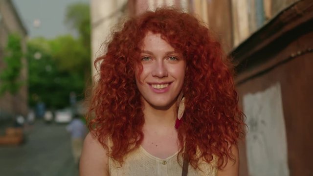 Happy young smiling woman with red curly hair look at camera walking in the city streets portrait slow motion summer face casual caucasian sunset beautiful lady outdoor closeup cute
