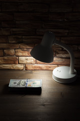 A lamp standing on a wooden table near a brick wall illuminates a bundle of dollars.