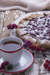 Fresh ripe juicy raspberries on baked pie with flowers and cup of tea on a wooden old background, decorated with a textile towel.