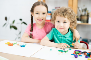Fototapeta premium Portrait of two happy kids smiling at camera while sitting at table in art and craft lesson of development school