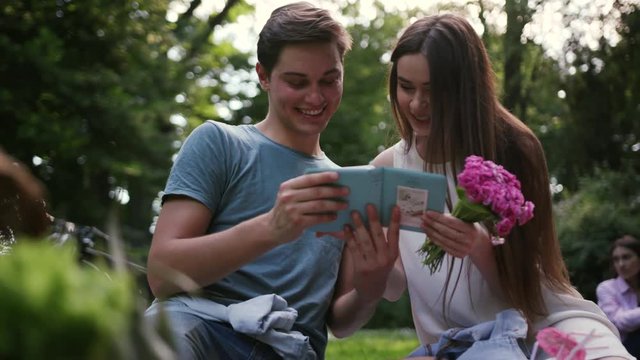 Portrait young couple see photo album smiling resting in park have picnic talk drink man eat apple woman love girl summer nature beautiful emotive outdoors outing pleasure positive pretty relations