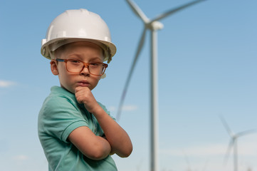 Serious smart little boy with glasses of white helmet stands against the background of windmills and the blue sky. Concept of dreams and future plans