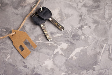 Two shiny metal keys with black plastic handle and rope tied to handmade house of brown goffred cardboard on old gray dirty cement floor with copy space. Estate concept