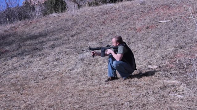Man kneeling on ry grass shooting an assualt rifle. 