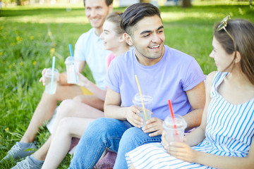 Cheerful teen couple with drinks talking at picnic on summer day with another couple on background