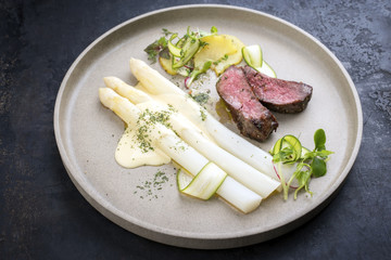 Modern barbecue dry aged sliced fillet steak with white asparagus in hollandaise sauce and roast potatoes as close up on a plate