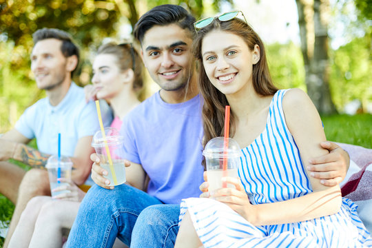 Young affectionate couple with drinks enjoying summer day in park with their friends on background