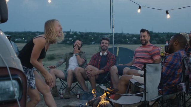 Group of diverse people chilling around bonfire in campsite and playing charades with woman acting and showing pantomime to others