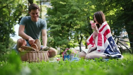 young woman with american flag resting read book in park have picnic talk drink man come give exotic fruit love girl summer beautiful outdoors outing pleasure positive pretty slow motion close up - Powered by Adobe