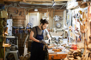 Young man in overalls standing by workbench and processing wooden workpiece