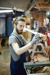 Young gloved worker looking at wooden workpiece before processing it in machine