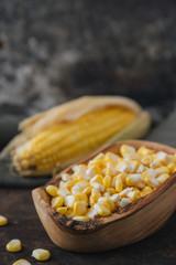 Corn kernels in a bowl next to a fresh corn on the cob on linen cloth over dark rustic background