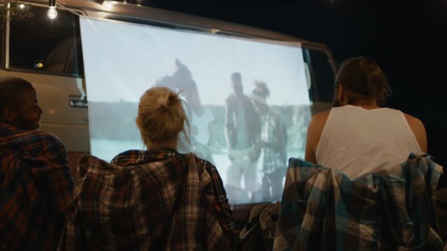 Group of friends having fun and relaxing while watching movie on screen on van in night time sitting around bonfire in camp
