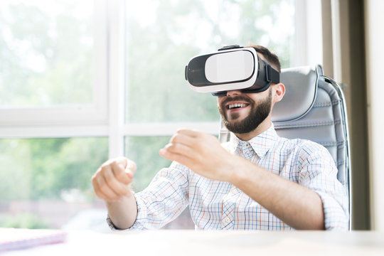 Portrait of handsome bearded businessman sitting at desk in office and enjoying VR simulator against window in sunlight, copy space