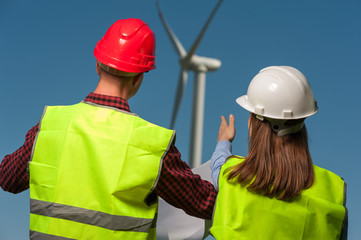 Rear view of two young engineers, a man and woman in helmets and in green waistcoats discussing a plan for designing against a windmill and a blue sky