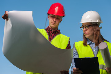Two young positive architects man and woman in green waistcoats evaluate work plan against blue sky...