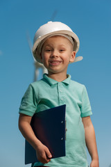 Portrait of a cheerful ambitious little boy with a helmet with an engineering plan in his hands against the backdrop of a windmill and a blue sky.