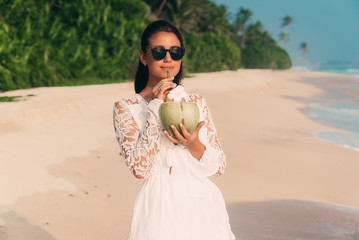 Fototapeta na wymiar Slender attractive dark-haired girl strolls along the seashore, wears a stylish tunic and sunglasses, drinks a delicious fresh coconut, smiles.