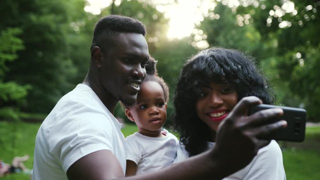 Happy african american family having fun in the park pose to take a selfie with mobile phone slow motion close up child girl daughter couple summer nature picnic mother parents portrait smartphone