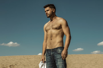 Strong face male model stripped wearing denim jeans and holding white t-shirt on the nature background with sand 