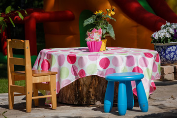 celebrations at home, outdoors, in the garden; on the table there is a colorful tablecloth with circles; at the table there is a wooden chair and a blue stool; On the table and next to the flower pots