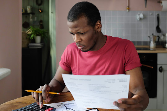 Indoor picture of young African American man sitting in his kitchen at home working with papers checking details in documents or bills calculating numbers with worried face looking troubled