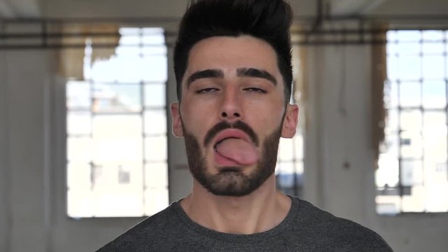 Young man joking, doing stupid expression with his tongue for the camera in a big empty room, acting silly