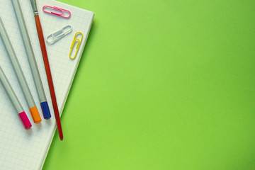 Colored pencils, paper clips and a brush on a notebook, green background with copy space