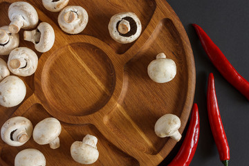 Still life of vegetables. A wooden plate on which contains champignons and pods of acute red pepper. Dark background