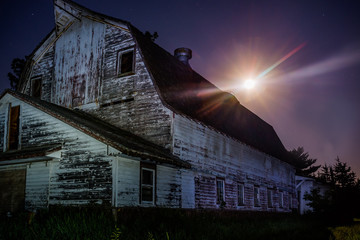 horizontal image old barn with full moon coming up behind.  Night shot using light painting to show abandoned barn and purple night sky with star burst effect on moon. 