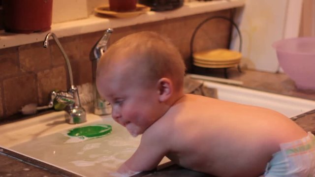 Small boy wearing only a diaper is playing in the overflowing kitchen sink and drinking the dish water.