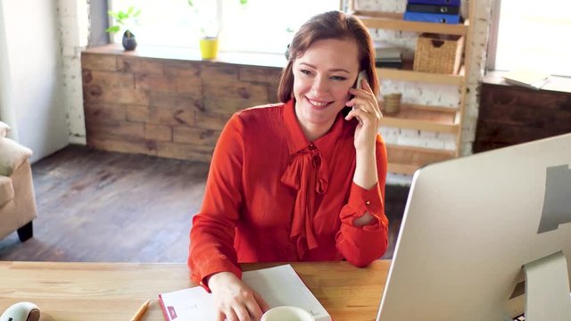 Sweet business lady recieving phone call in office. Lovely businesswoman wearing formal clothes talking on phone with genuine smile and then drinking coffee. Modern office interior.