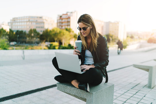 Photo of a freelancer female enjoying take away coffee while working outdoors on a portable computer connected to public wifi. Stylish student girl studying online while spending free time in a park