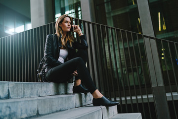 Charming caucasian woman with long blonde hair wearing stylish leather jacket talking by a mobile phone while sittig on the steps of a street staircase on a blurred business building background.