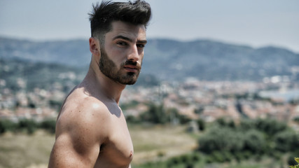 Handsome shirtless muscular young man outdoor on a balcony or terrace, wearing only shorts, looking at camera