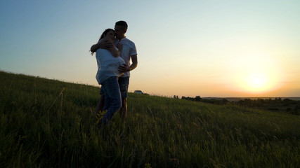Fototapeta premium Romantic young happy pregnant couple hugging in nature at sunset. Man hugging woman from back in the evening in fields. Family, marriage, love, devotion, faith concept