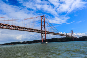Bridge Ponte 25 de Abril in Lisbon in front of a blue sky with some clouds over the sea.