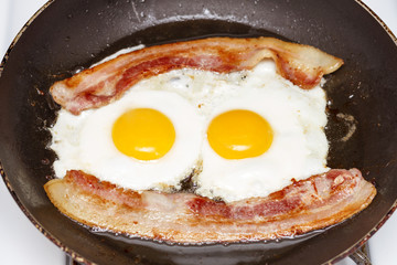 Smiling Happy Face Frying Eggs breakfast with bacon in iron vintage pot.