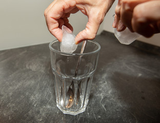 Hands are putting ice cubes into a long glass with a long spoon.