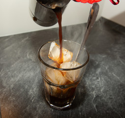 A person is pouring coffee from a coffee machine into glass with filled with ice cubes and a long spoon.