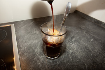 A person is pouring coffee from a coffee machine into glass with filled with ice cubes and a long spoon.