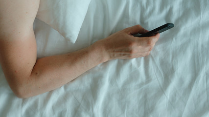 TOP VIEW: Human hand with a phone on a bed in the morning
