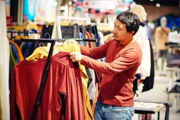 Smart man with beard choosing clothes in clothing store at shopping center, looking for new shirts design that hanging on the rail, Fashion and Consumerism Concept.