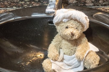 Bear wears towel on head and body with tissue paper is in toilet.