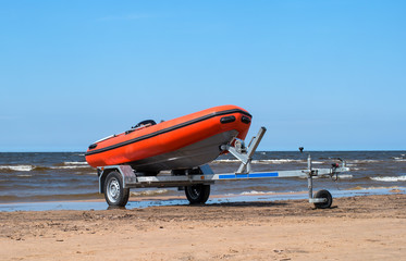 Lifeboat on trailer. The lifeboat is orange. The trailer with the boat is on the shore by the water. Sandy beach