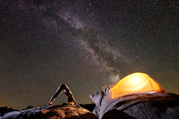 Attractive girl doing gymnastic yoga exercises on big boulder on dark starry evening sky with Milky way, brightly lit tourist tent and black mountains background. Active lifestyle, camping concept.
