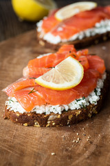 Tasty salmon sandwich with cream cheese and slice of lemon on wooden board, closeup view, selective focus