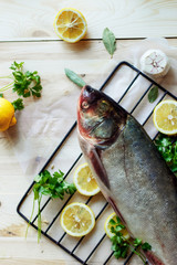 Fresh uncooked silver carp with lemon, herbs and spices on parchment over wooden board, top view