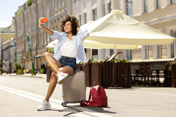 Selfie time. Cute curly girl taking selfies while sitting on her suitcase in the middle of the...